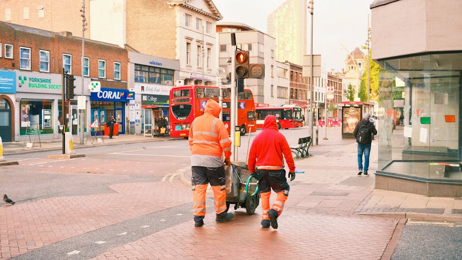 A sanitation worker dressed in bright orange high-visibility uniform with reflective stripes, standing outdoors on a paved surface near a modern building with glass windows, holding a broom in the rig