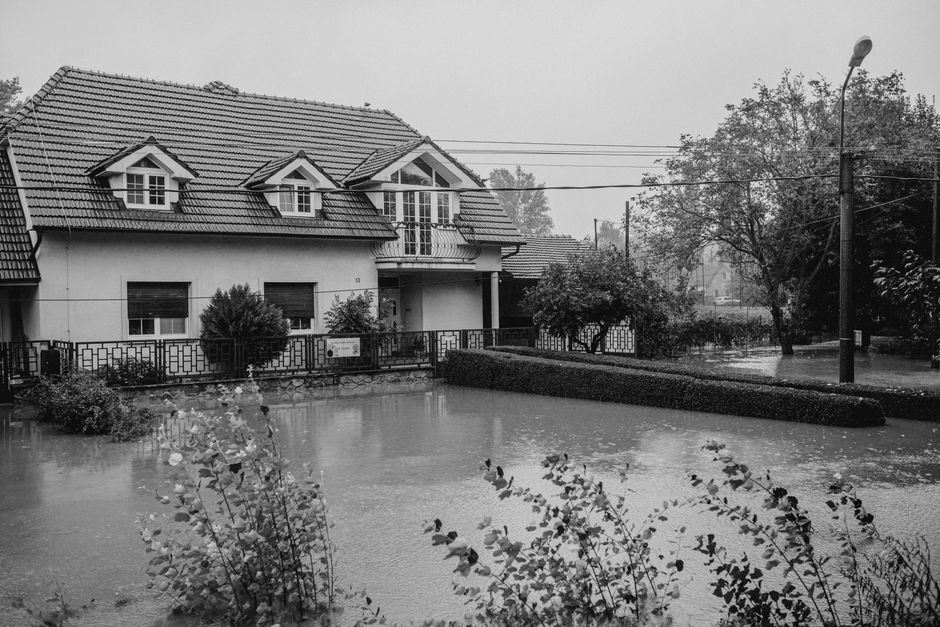 A residential area during a heavy flood, showcasing a house with a tiled roof and multiple dormer windows, surrounded by flooded gardens and streets. Water covers the driveways, lawn, and parts of the