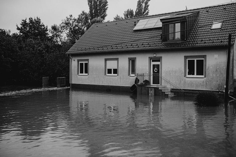 A residential area during a heavy flood, showcasing a house with a tiled roof and multiple dormer windows, surrounded by flooded gardens and streets. Water covers the driveways, lawn, and parts of the