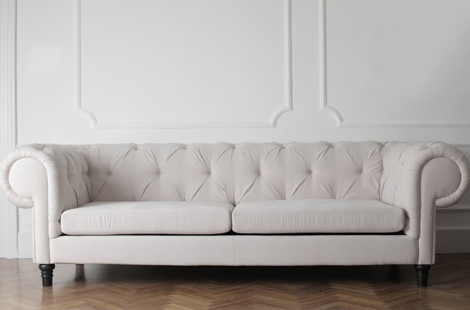 A bright, modern living room with a light grey fabric sofa adorned with matching and darker grey cushions, placed on a beige carpet. Behind the sofa, large floor-to-ceiling windows with sheer white cu